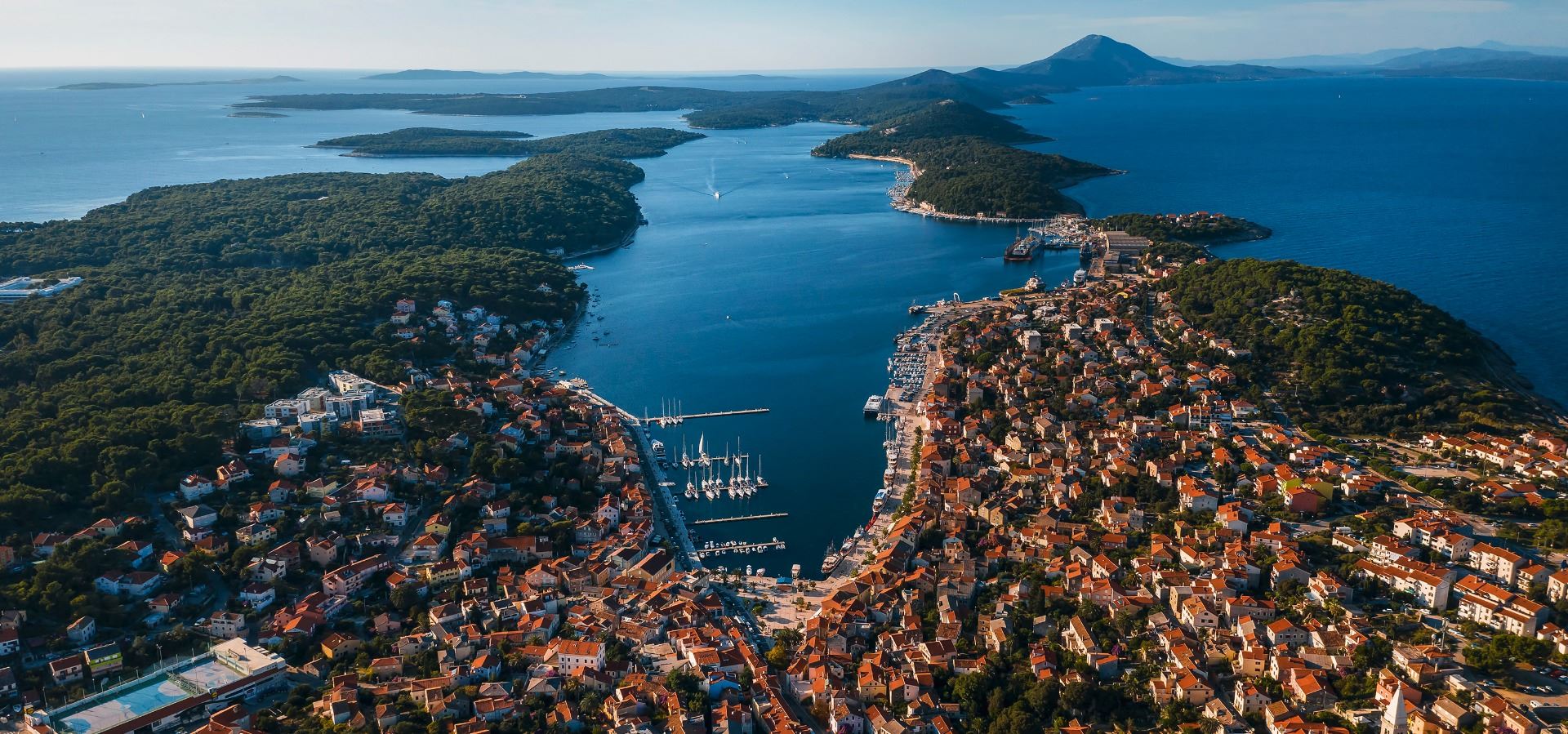 Copyright: Ivan Brcic, visitmali-losinj.croatia.hr An aerial view of Mali Lošinj, Croatia, showcasing the vibrant coastal town nestled around a deep, sheltered bay filled with sailboats. Lush green islands and the open sea stretch into the distance under a clear sky.
