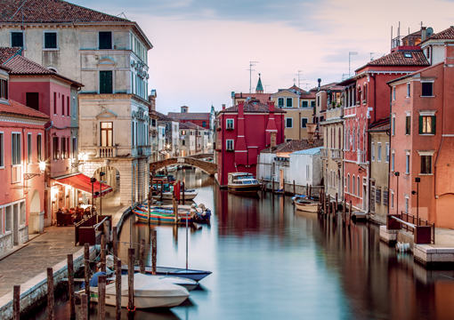 A tranquil canal scene in Chioggia, Italy, showcasing the town's vibrant colors and historic architecture. Calm water reflects the pastel-colored buildings lining the canal, with small boats moored along the sides.
