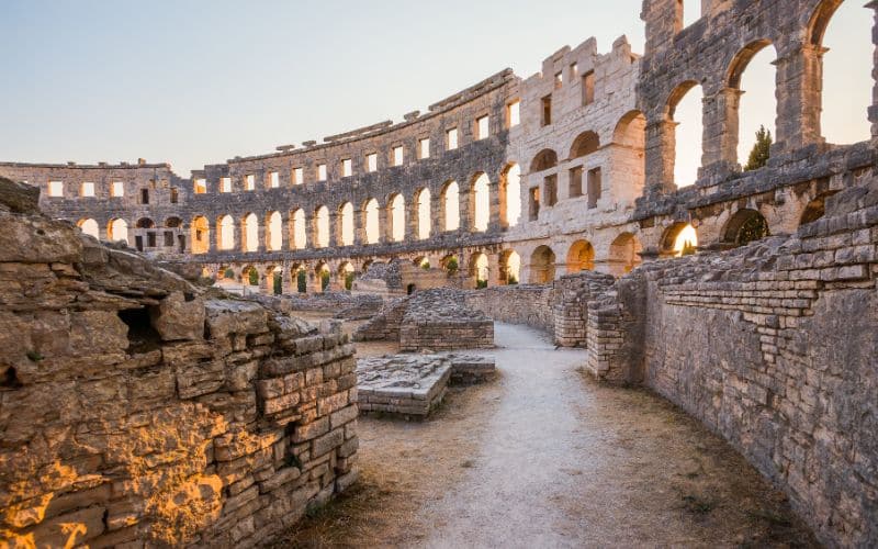 Interior view of the Pula Arena, a well-preserved Roman amphitheater in Pula, Croatia. Sunlight streams through arched openings along the upper walls, illuminating the stone structure.
