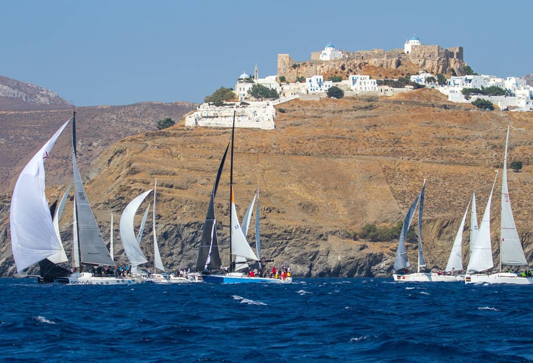 Sailboats racing in the Aegean 600 regatta on deep blue waters, passing below the whitewashed village and Venetian castle of Astypalaia perched on a rocky hilltop.