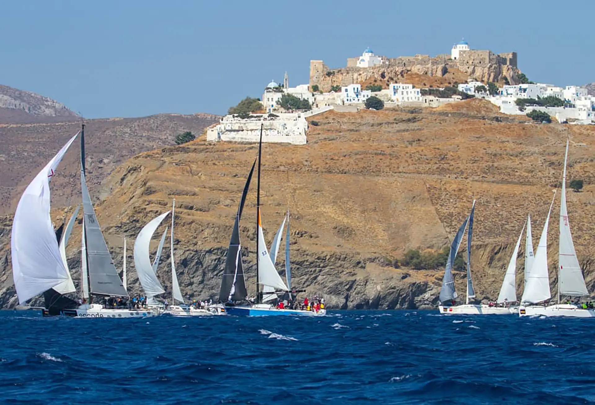 Sailboats racing in the Aegean 600 regatta on deep blue waters, passing below the whitewashed village and Venetian castle of Astypalaia perched on a rocky hilltop.