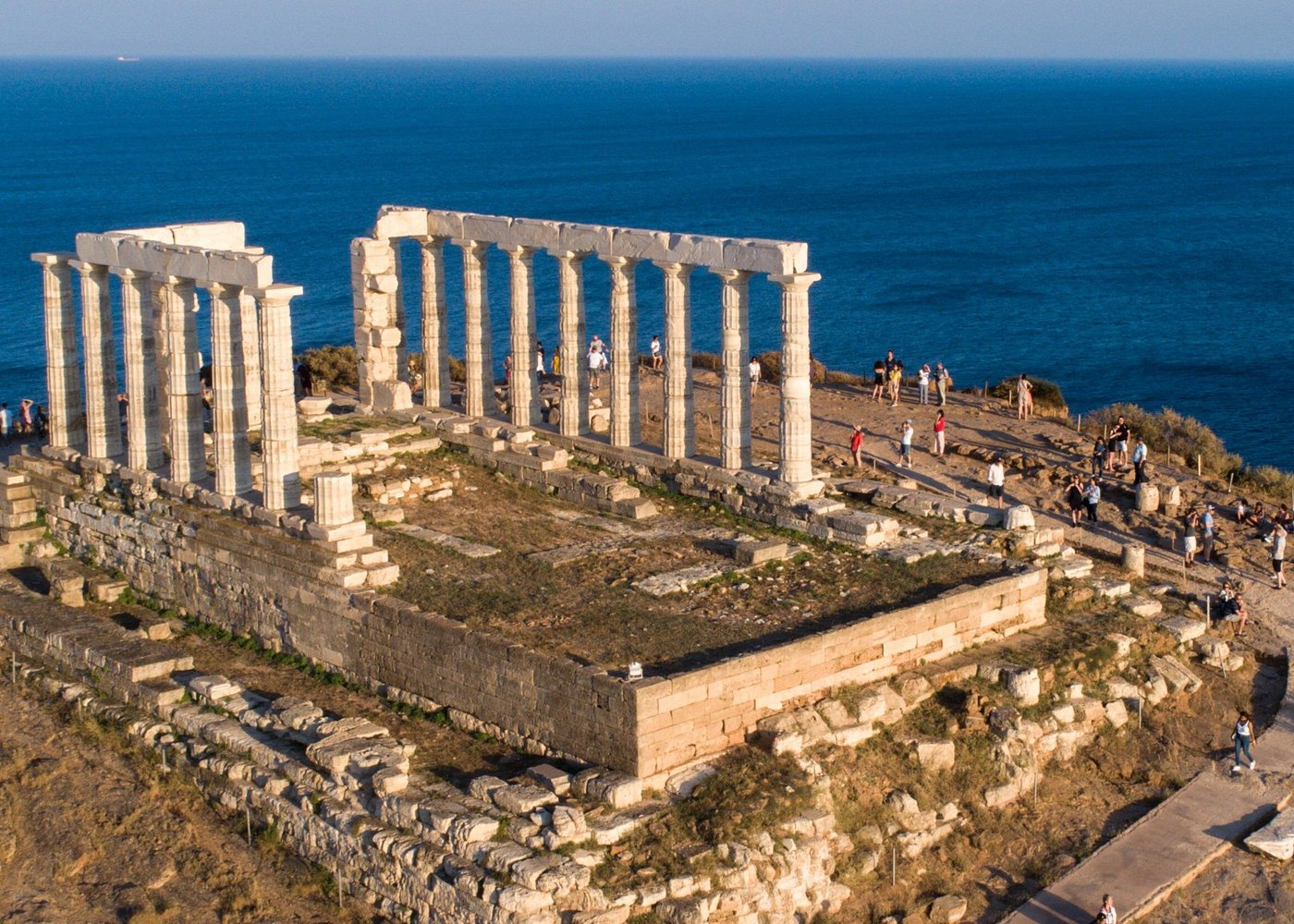 High-angle photograph of the Temple of Poseidon ruins at Cape Sounion, displaying ancient white Doric columns atop a rocky promontory overlooking the deep blue Aegean Sea, with small figures of tourists exploring the site.