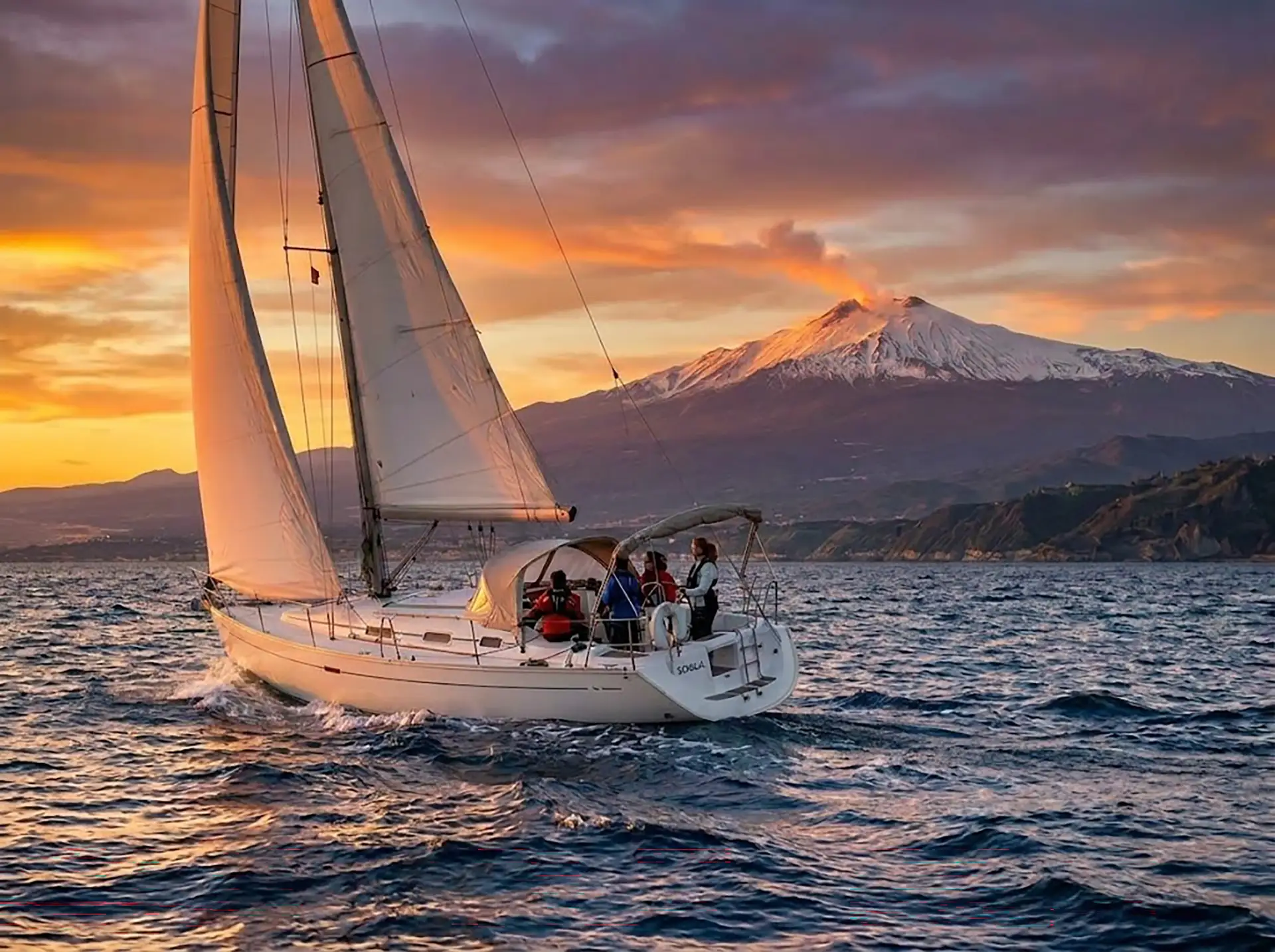 Yacht under sail at sunset with snow-capped Mount Etna rising on the Sicilian horizon