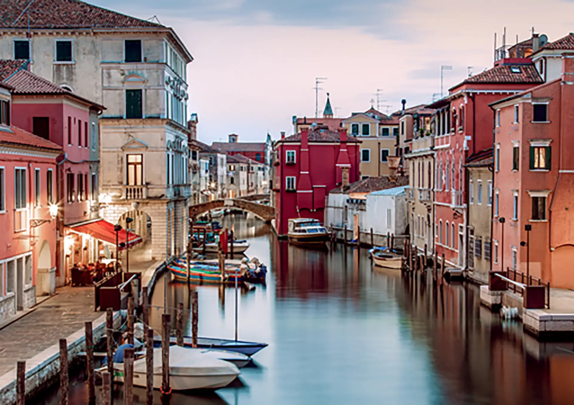 A tranquil canal scene in Chioggia, Italy, showcasing the town's vibrant colors and historic architecture. Calm water reflects the pastel-colored buildings lining the canal, with small boats moored along the sides.