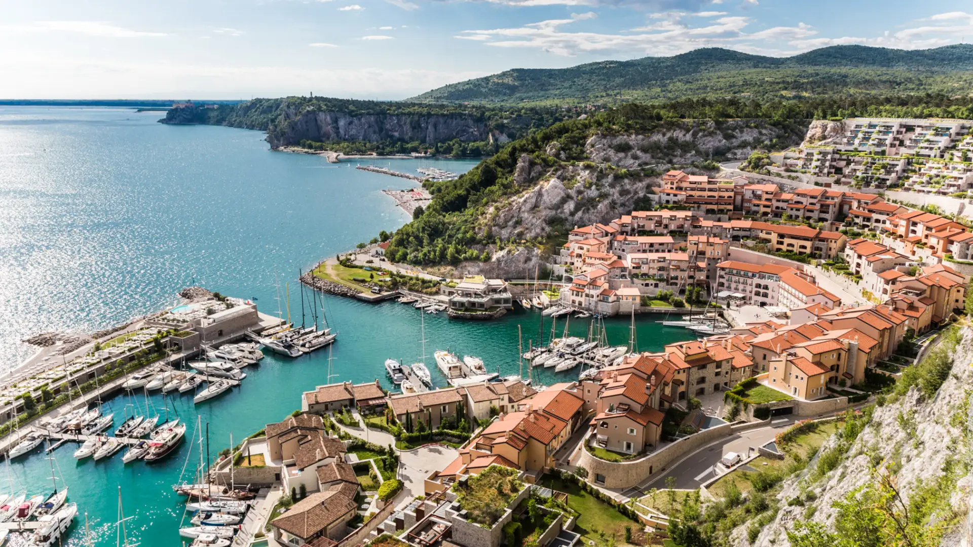 Aerial view of Portopiccolo, a modern eco-resort in Italy, blending terracotta-roofed buildings into a cliffside setting overlooking the turquoise Adriatic Sea.