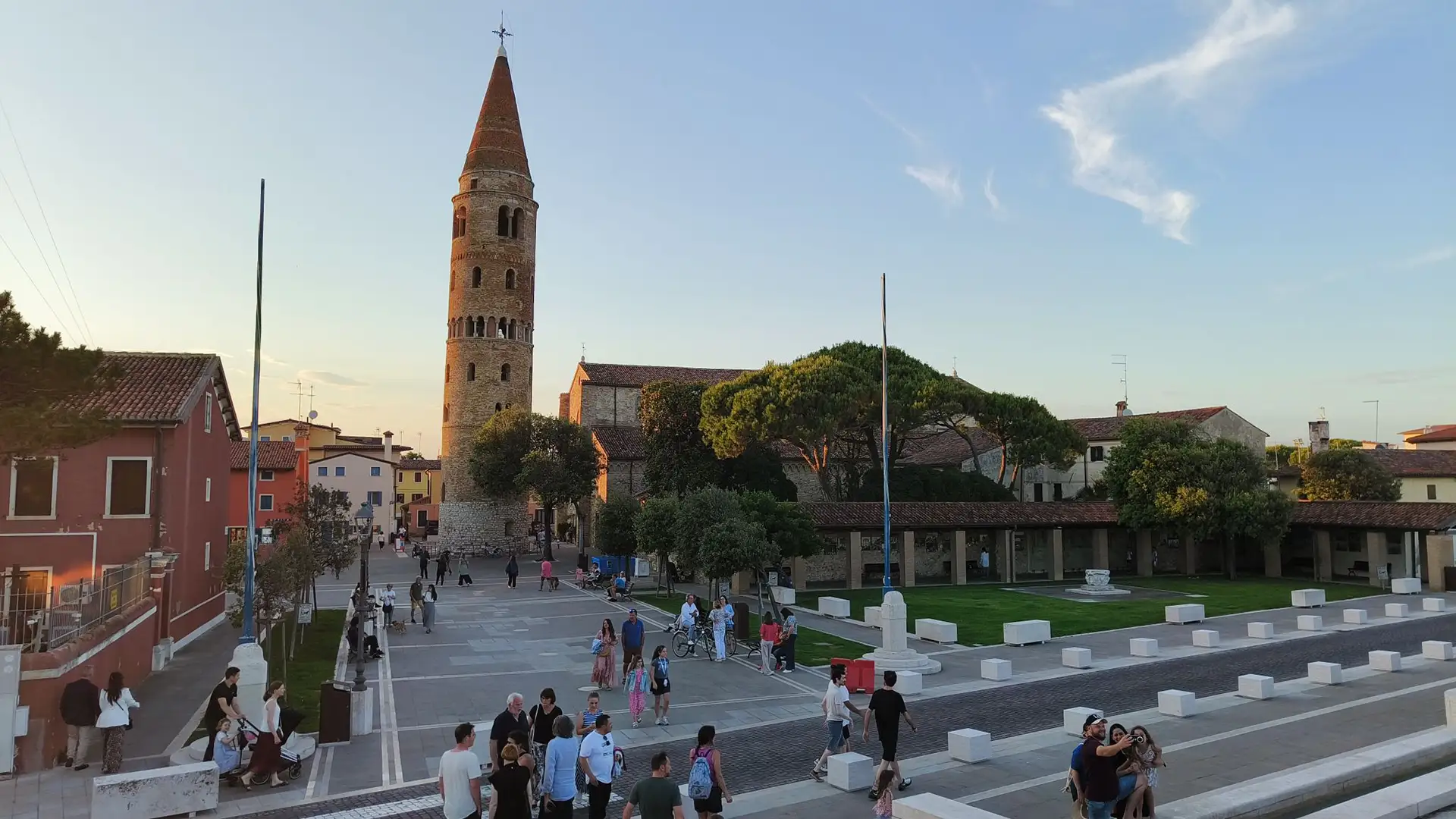 The iconic cylindrical bell tower of Caorle's Duomo di Santo Stefano standing tall in the late afternoon sun, overlooking a busy town square with people walking.
