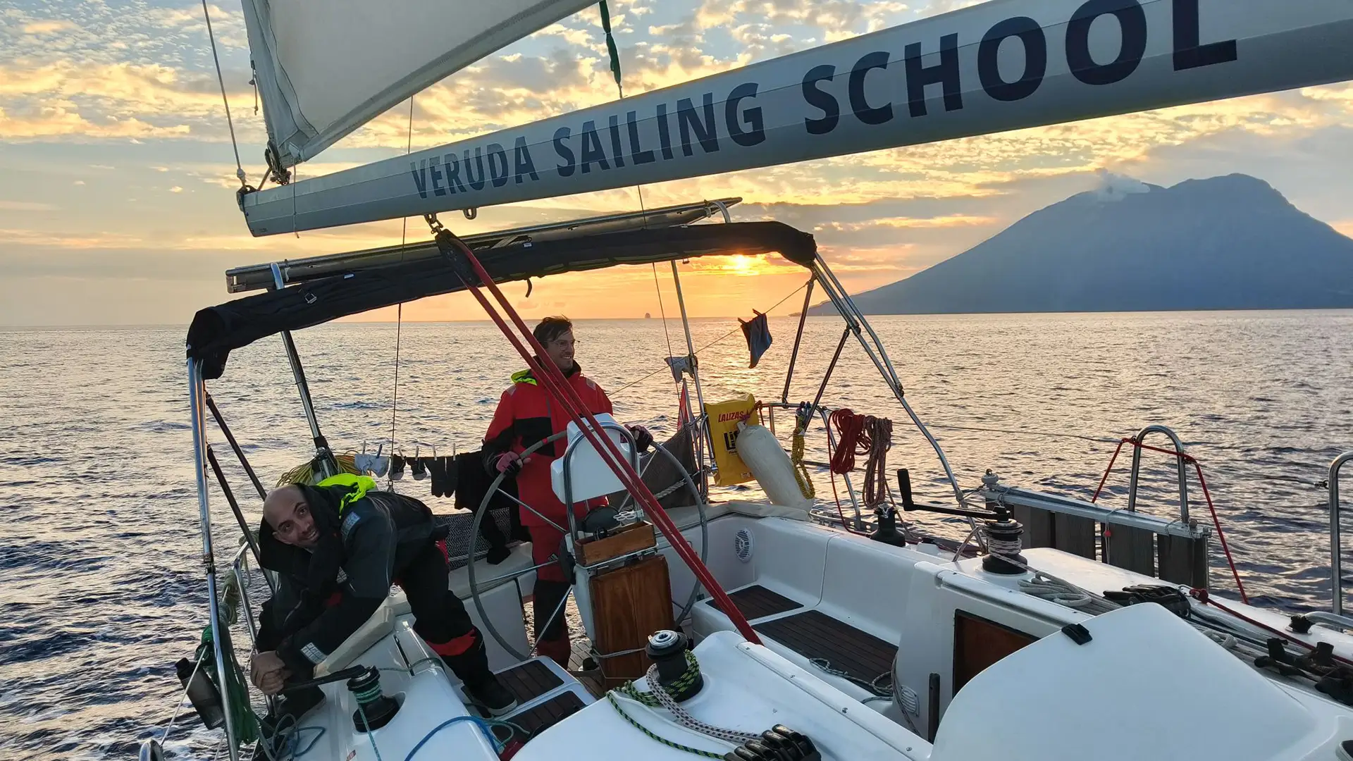 A racing crew on a offshore sailboat. Behid the boat, the large volcanic cone of Stromboli volcano is visible, spewing clouds of steam.