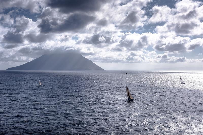 Copyright: Rolex / Kurt Arrigo An offshore sailboat race with multiple yachts on a choppy sea. In the distance, the large volcanic cone of Mount Etna is visible, partially obscured by clouds.